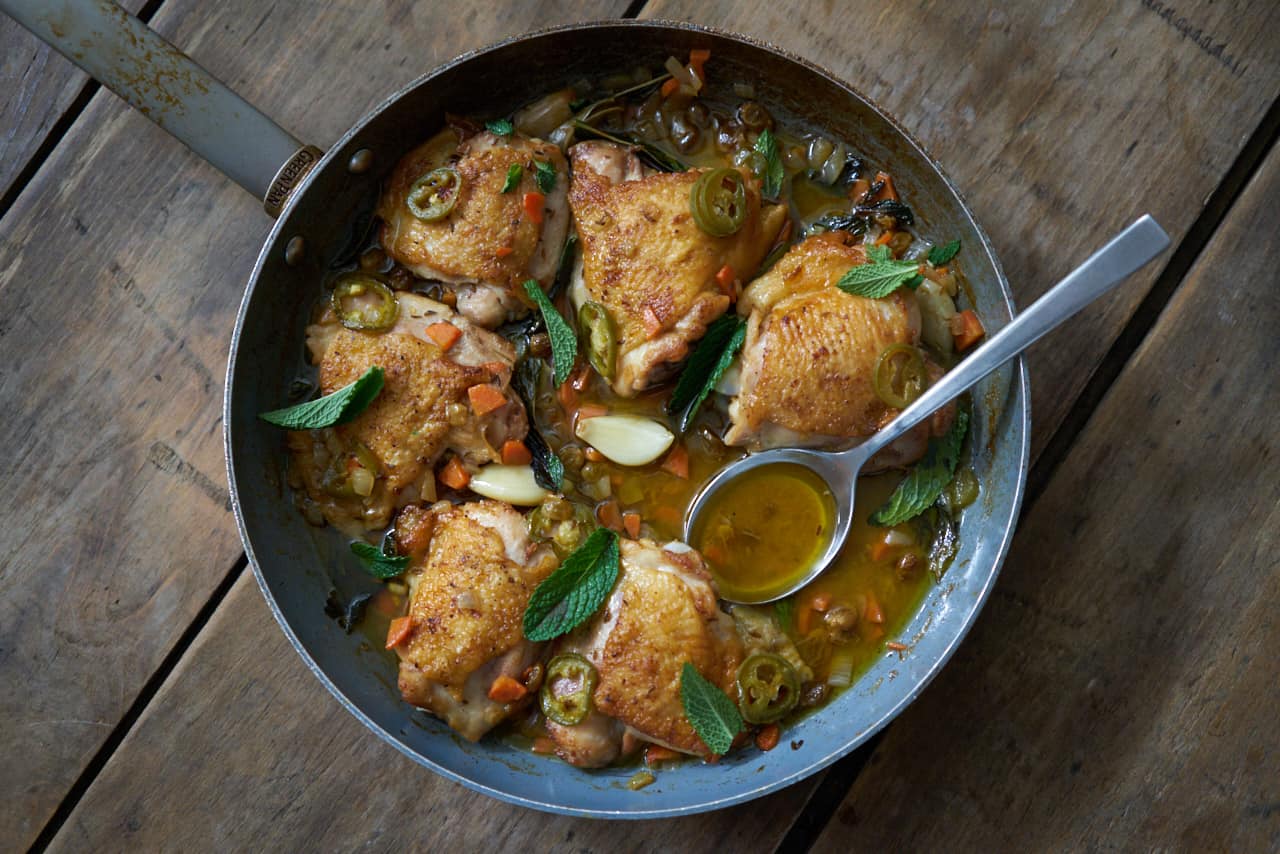 A skillet with chicken escabeche sits on a wooden surface. A sliver spoon is in the pan on the right side. Garlic, golden raisins and jalapeños can be seen in the escabeche sauce, and the dish is garnished with fresh mint.