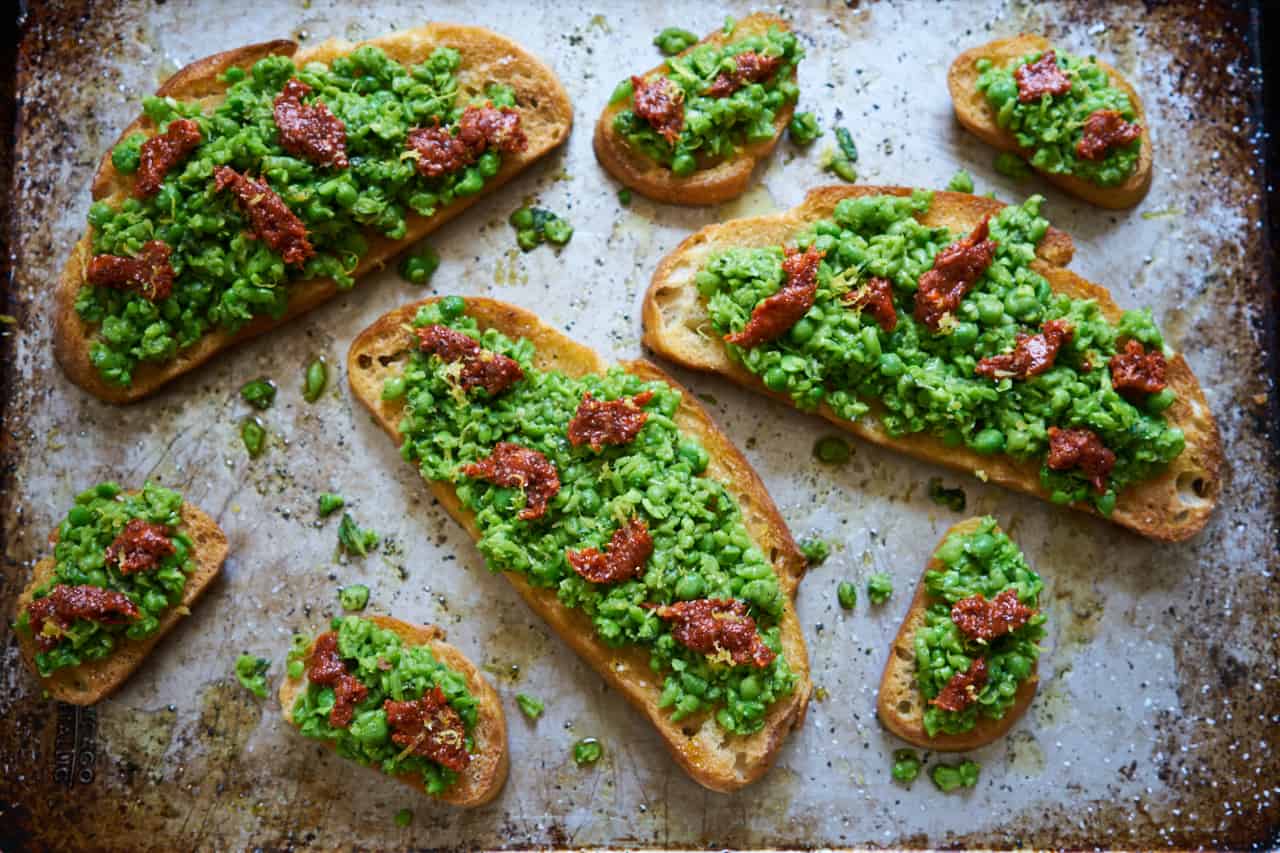 Tartines and crostini with fresh spring peas mixed with mint and lemon and topped with nduja are displayed on a baking sheet.