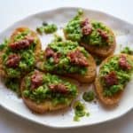 Five pea crostini topped with n'duja on a white plate.