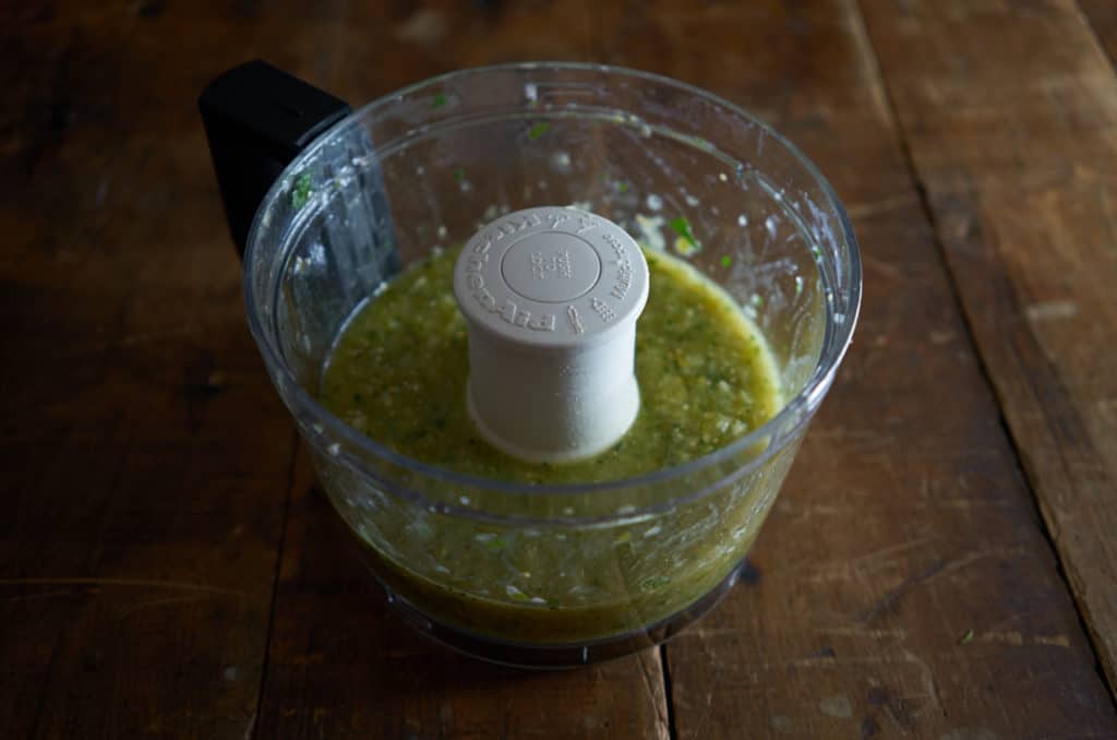 A food processor bowl containing the blended ingredients: cooked tomatillos, serrano chiles, onions and cilantro.