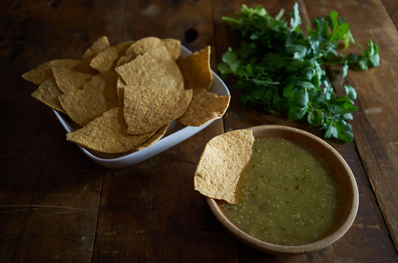 A wooden bowl of salsa verde with a tortilla chip in it, alongside a bowl of tortilla chips and a bunch of fresh cilantro.