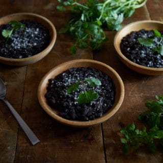 3 wooden bowls of Mexican black beans, a sliced avocado, and fresh cilantro on a wooden table.
