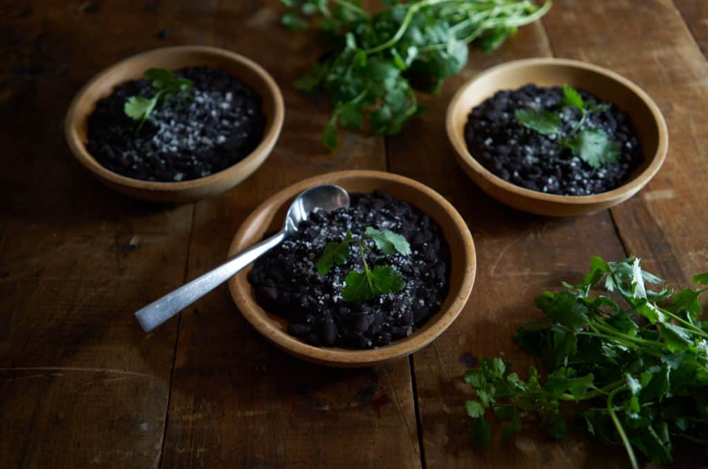 3 wooden bowls of Mexican black beans and fresh cilantro on a wooden table.