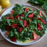 A plate of salad with arugula, herbs, and sliced strawberries on a brown surface with basil leaves and lemons in the background.
