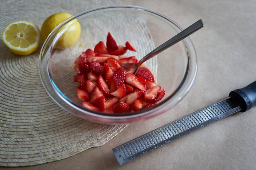 A glass bowl filled with sliced strawberries and a silver-colored spoon sits on a woven placemat. A zester is in the foreground and lemons are in the background.