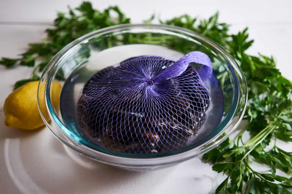 PEI mussels in a purple net sit in a glass bowl filled with water. Parsley and a lemon are in the background on a white surface.