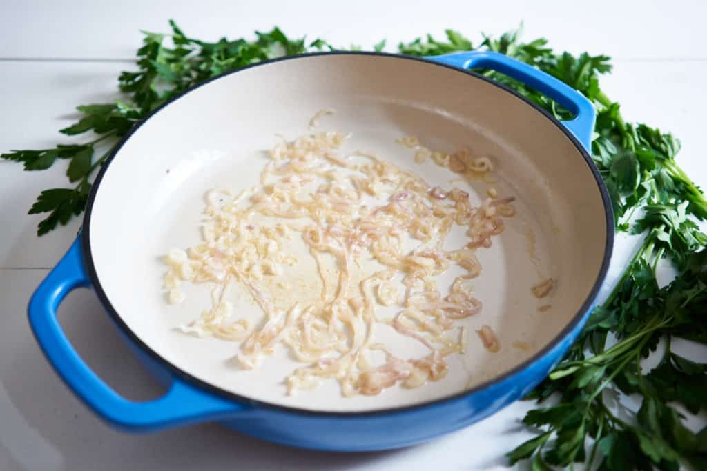 Cooked shallots in a blue casserole dish with a cream-colored interior. Parsley surrounds the dish on a white surface.