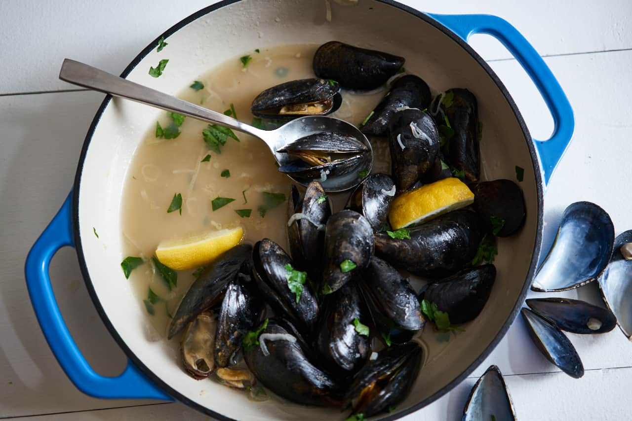 A blue casserole dish with mussels in white wine sauce (moules marinières) and lemon wedges sits on a white surface with empty mussel shells to the right of the dish.