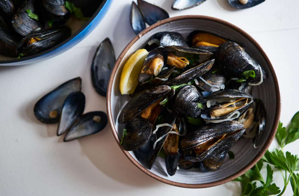 A bowl of mussels in white wine (moules marinières) on a white surface surrounded by parsley and empty mussel shells. A blue bowl of cooked mussels is in the upper left corner.