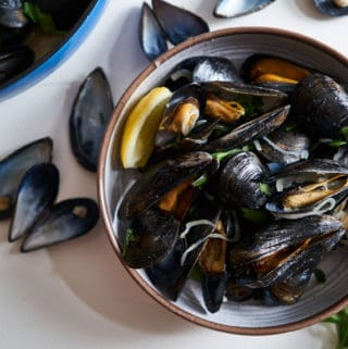 A bowl of mussels in white wine (moules marinières) on a white surface surrounded by parsley and empty mussel shells. A blue bowl of cooked mussels is in the upper left corner.