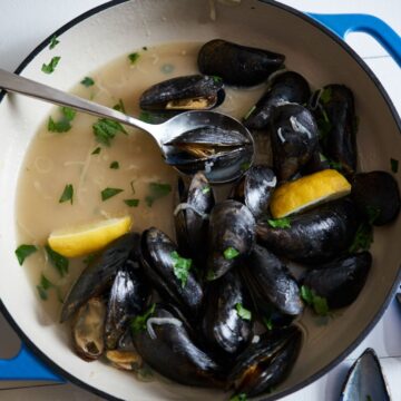 Mussels in white wine sauce with lemon wedges and parsley in a blue pan with a spoon in it.