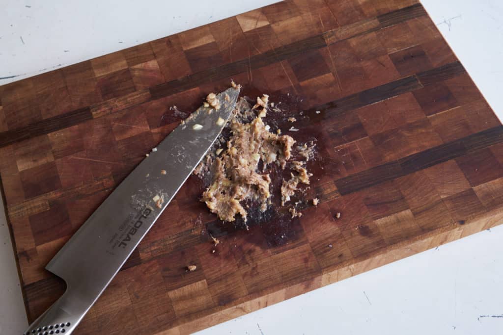 A large chef's knife sits next to anchovy and garlic that have been smeared together to form a paste on a wooden cutting board.