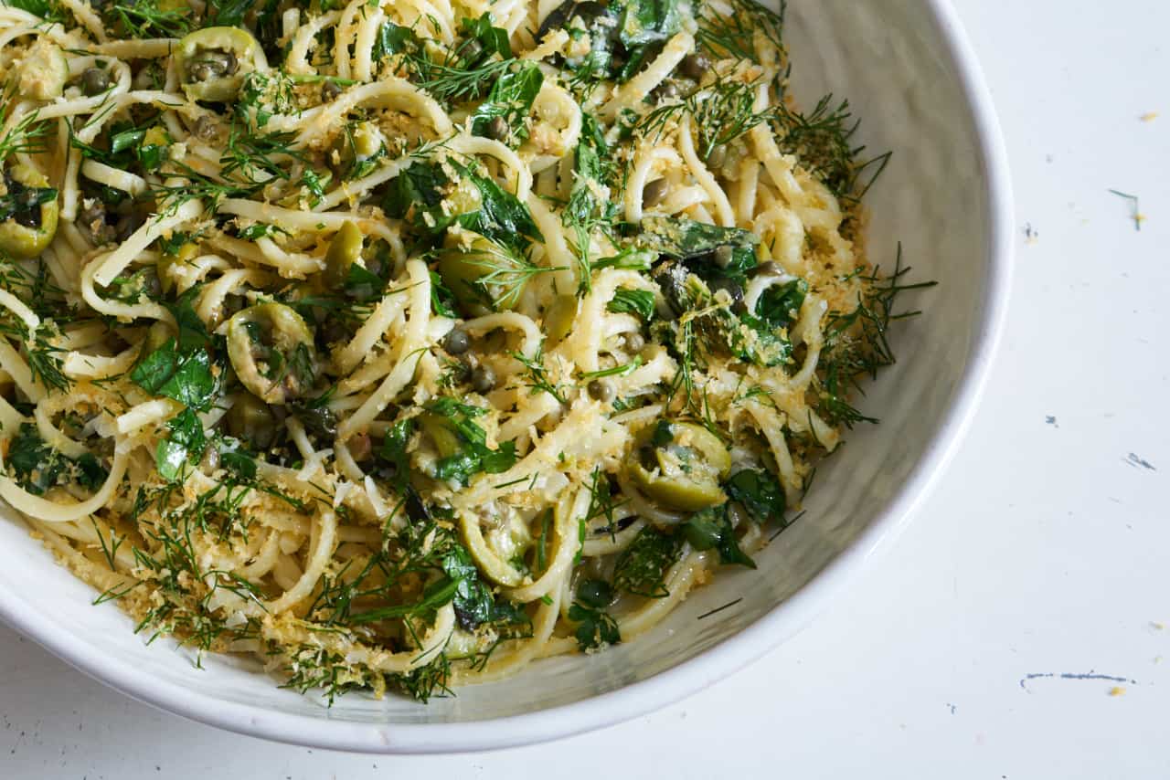Linguine pasta with green olives, herbs, and capers topped with bread crumbs in a white bowl sitting on a white surface.