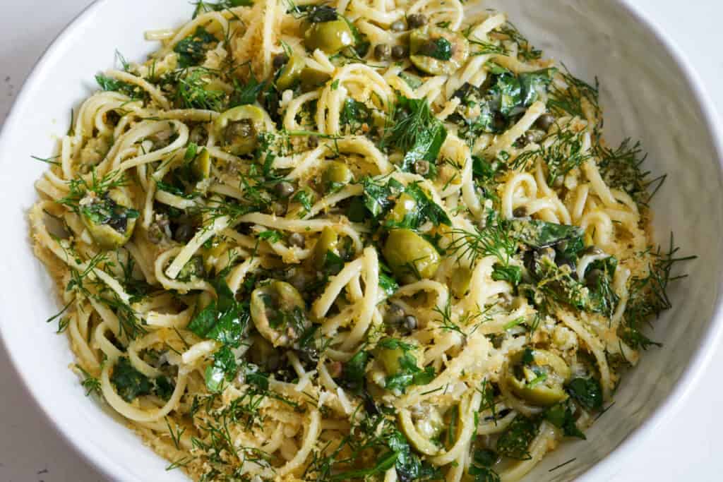 Linguine pasta with green olives, herbs, and capers topped with bread crumbs in a white bowl sitting on a white surface.