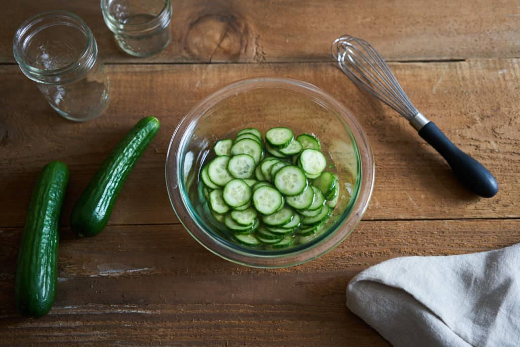 Pickled Persian cucumbers in a glass bowl on a wooden surface. A whole cucumber is on the left, a whisk is on the right.