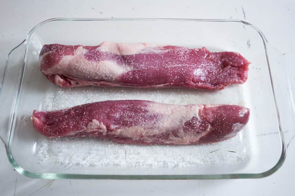 Two salted raw pork tenderloins in a glass casserole dish on a white surface.