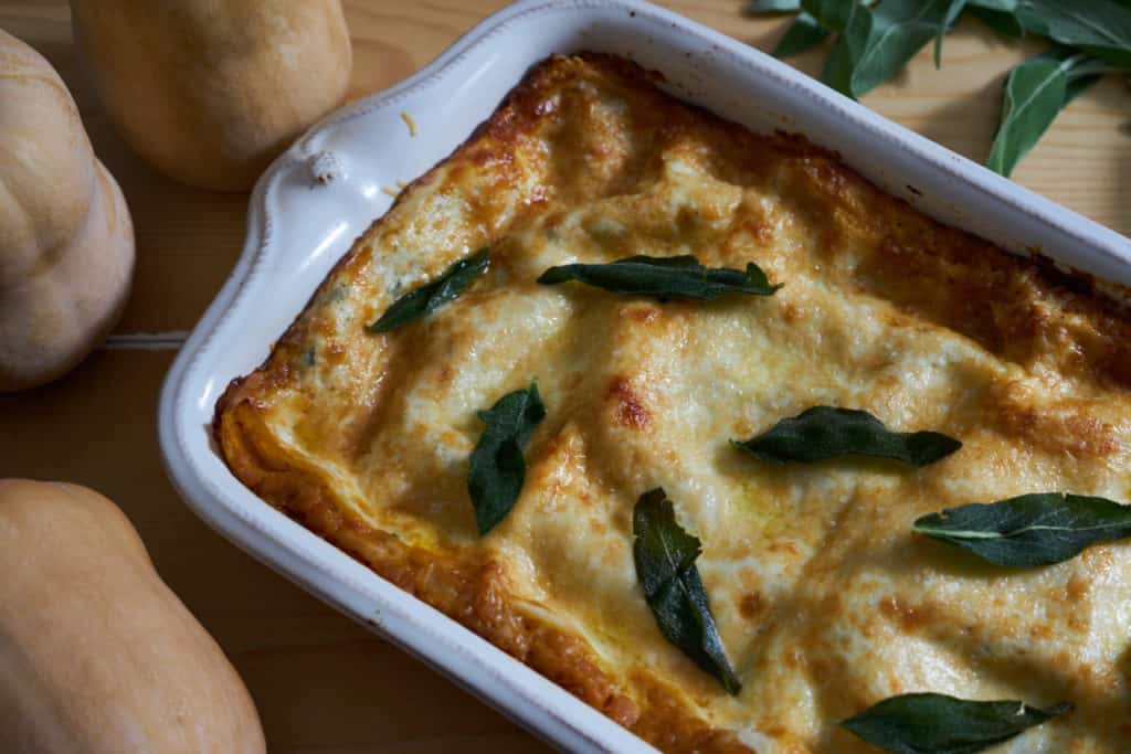 A white baking dish with butternut squash lasagna topped with fried sage surrounded by fresh sage and whole butternut squash.