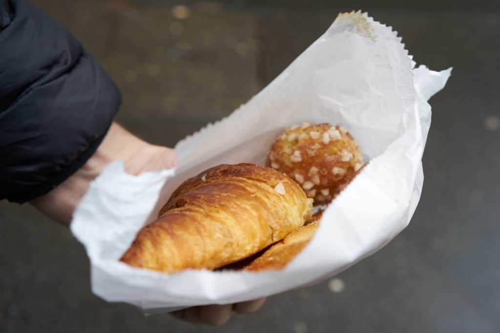 A man's hand is shown holding a white paper bag with a croissant, a chouquette, and a palmier. 