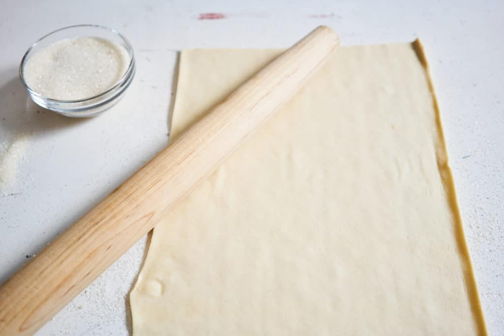 A sheet of puff pastry, a wooden rolling pin, and a small bowl of sugar.