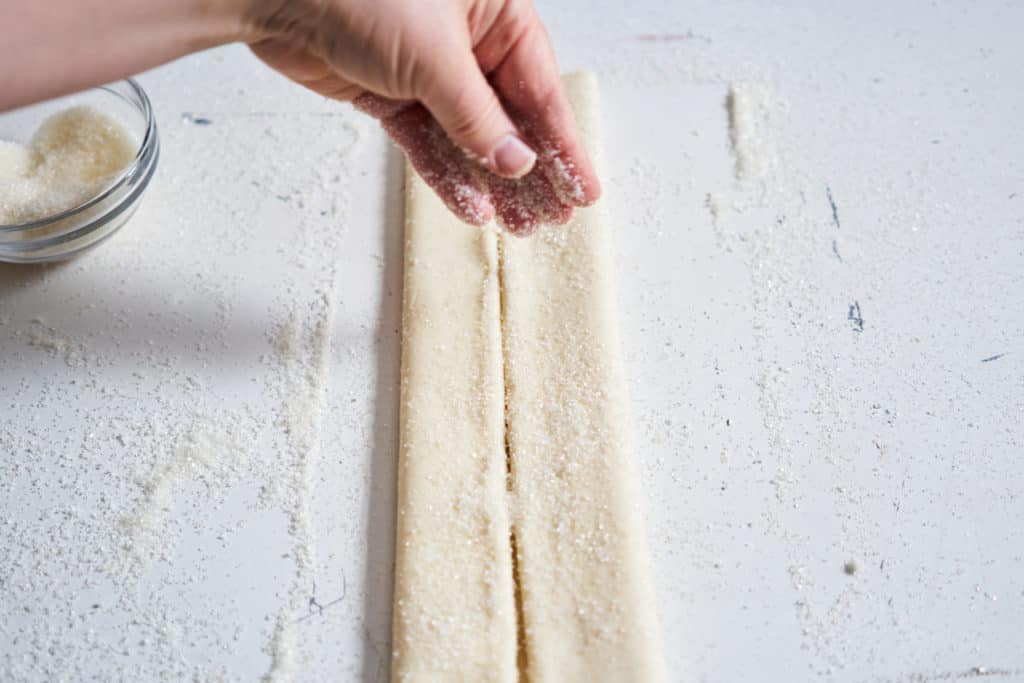 A woman's hand is sprinkling sugar onto a sheet of puff pastry that has been folded over itself twice.