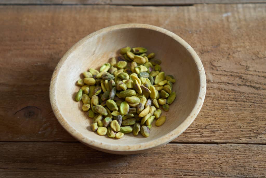 A small wooden bowl of raw pistachios on a wooden surface.
