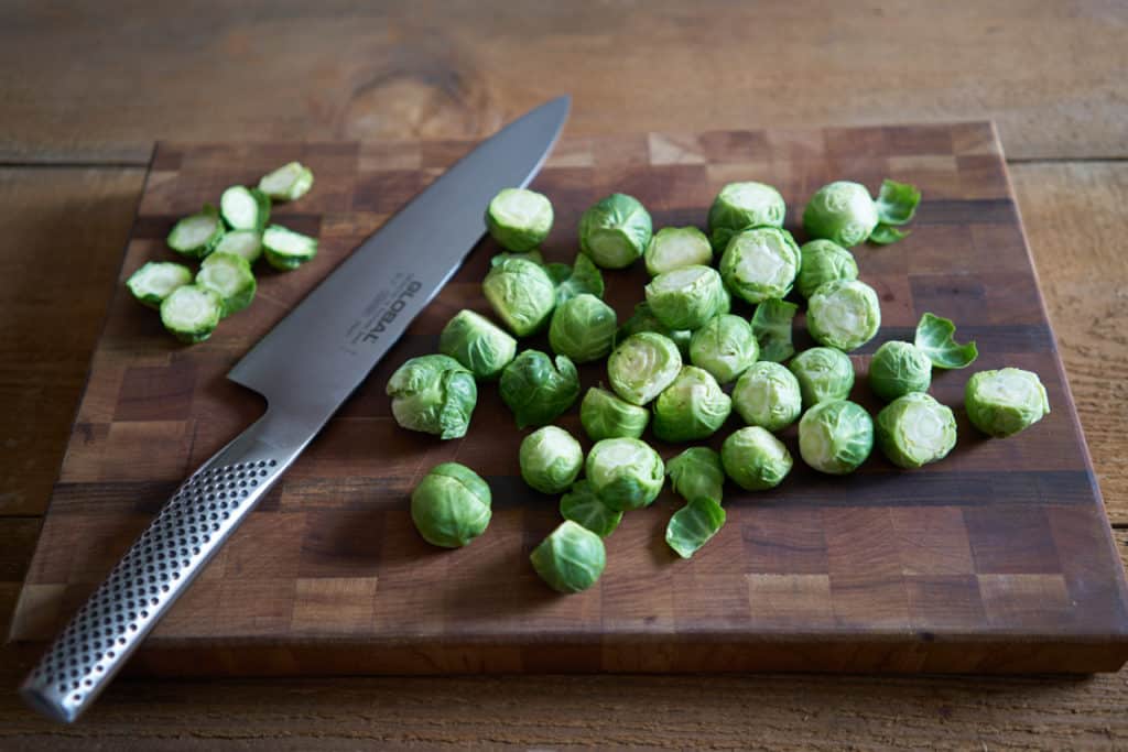 A wooden cutting board with trimmed brussels sprouts and a chefs knife.