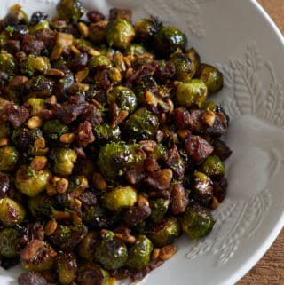 Roasted brussels sprouts with pistachios, dates, and lime in a white serving bowl on a wooden surface.