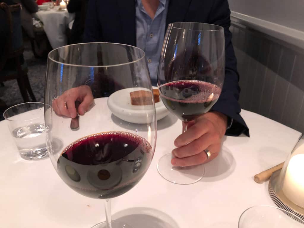 Two large glasses of red wine on a white tablecloth. A man's hand is holding the stem of one of the glasses.