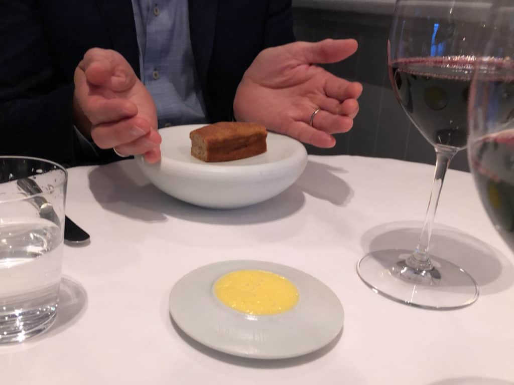 A man's hands are cuppping a small loaf of gluten-free cornbrread on a white plate. A small dish of butter and glasses of red wine are also on the table.