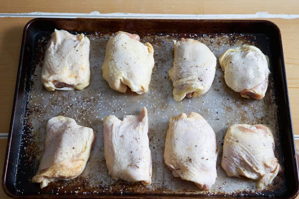 Raw chicken thighs seasoned with salt and pepper on a baking sheet.