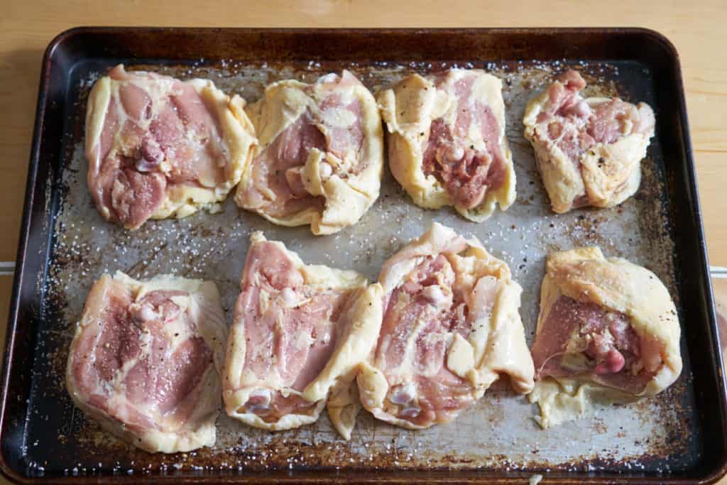 Raw chicken thighs seasoned with salt and pepper on a baking sheet.