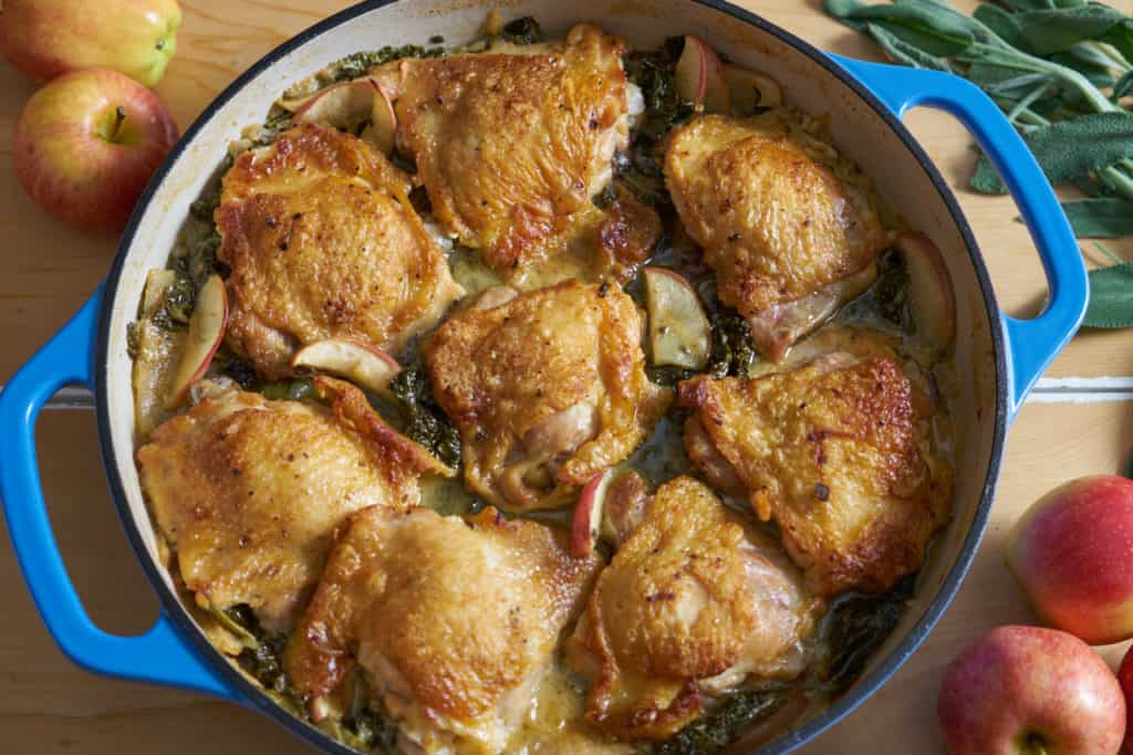 A blue casserole dish containing dijon chicken thighs with apples and kale is on a wooden surface, surrounded by whole apples and fresh sage leaves.