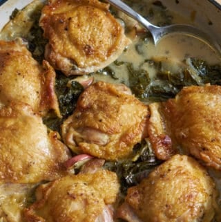 A blue casserole dish containing dijon chicken thighs with apples and kale is on a wooden surface with a silver spoon resting in the sauce.