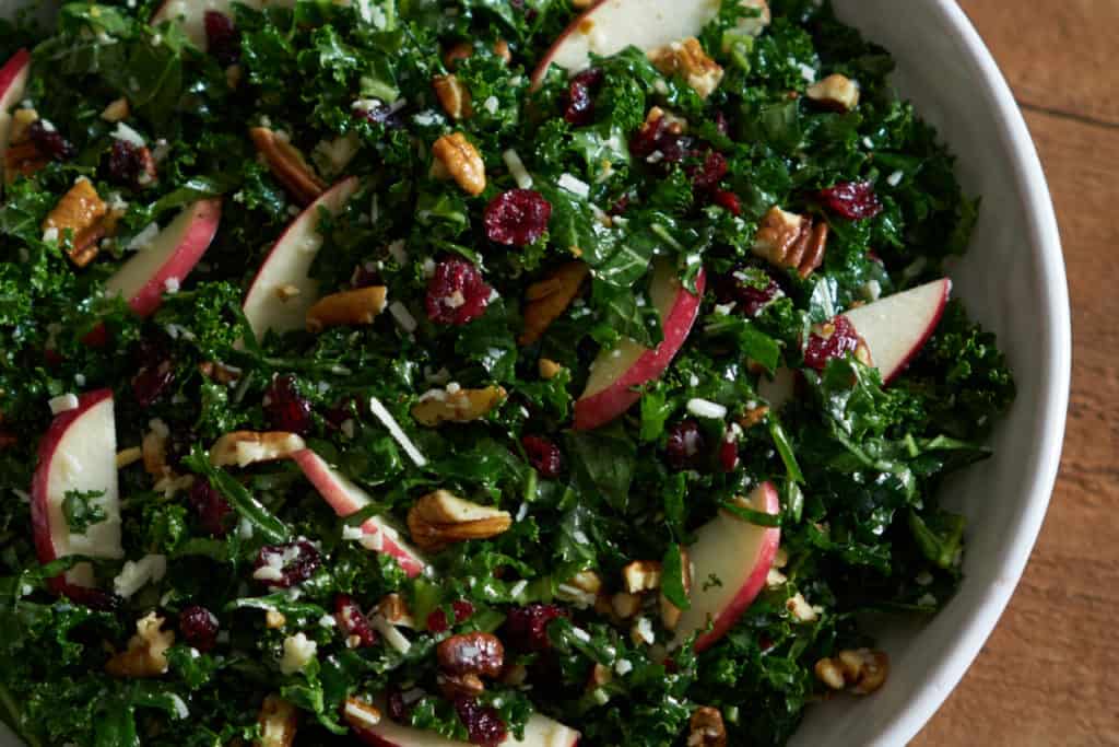 Kale salad with cranberries, apples and pecans in a white bowl on a wooden surface.