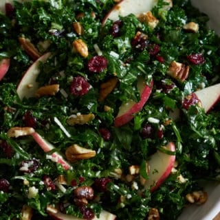 Kale salad with cranberries, apples and pecans in a white bowl on a wooden surface.