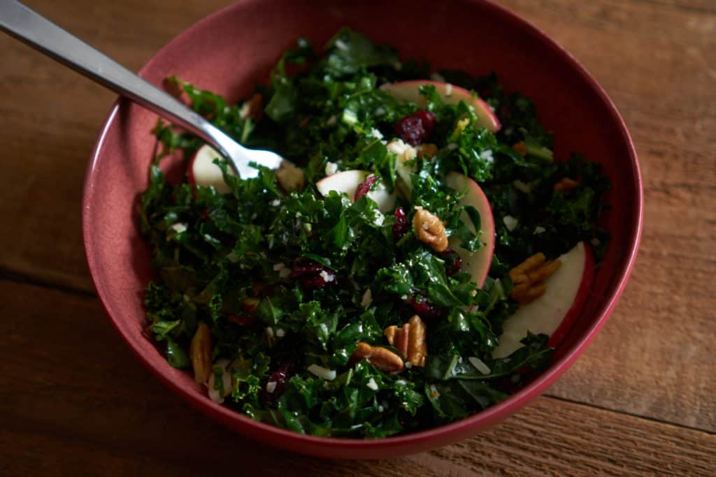 Massaged kale salad with cranberries, apples and pecans in a red bowl with a fork in it on a wooden surface.