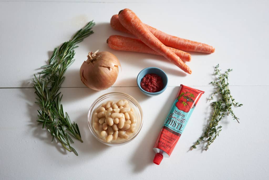 Fresh herbs, an onion, carrots, cannellini beans and a tube of tomato paste on a white surface.