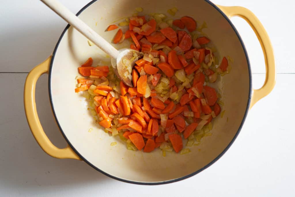 A wooden spoon rests in a yellow pot of sautéed carrots and onions.