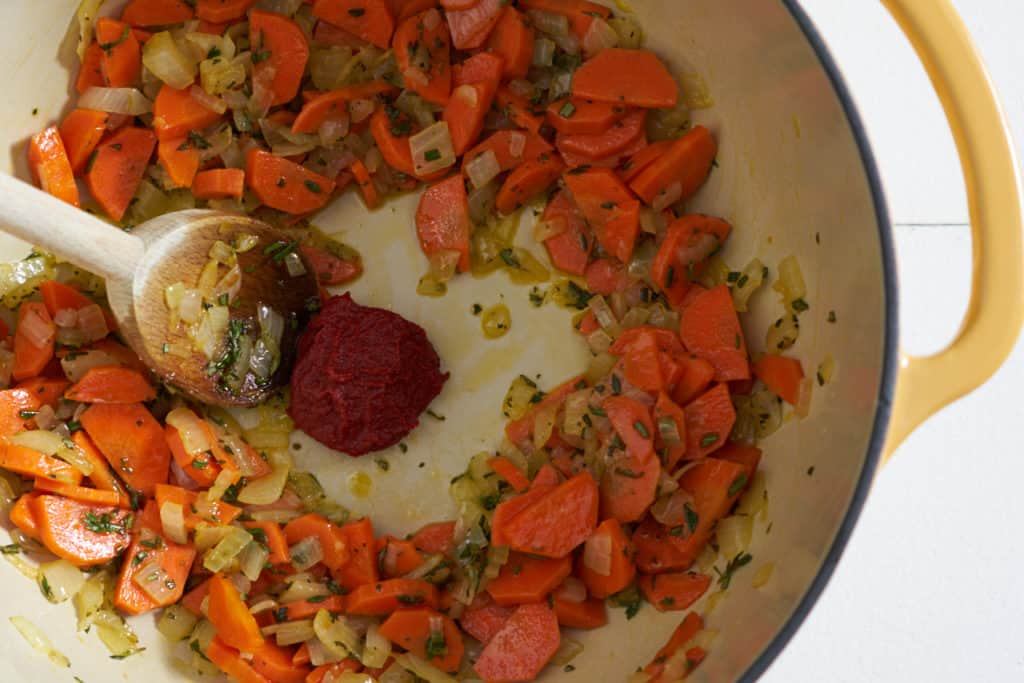 Tomato paste in the center of a yellow pot with sautéed carrots, onions, and herbs.