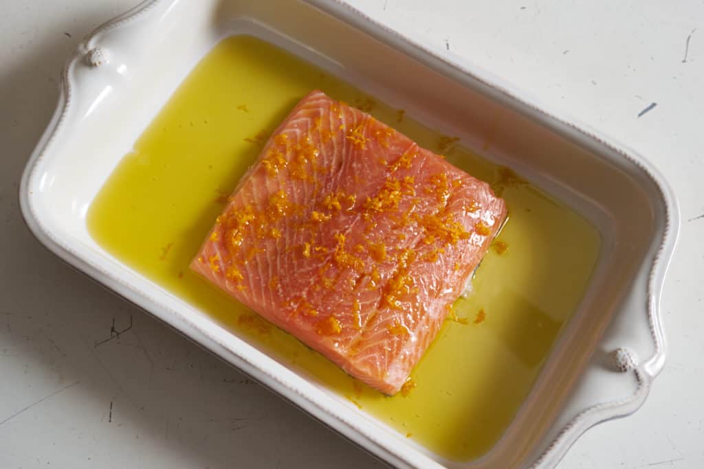 A salmon filet topped with citrus zest surrounded by olive oil in a white baking dish.