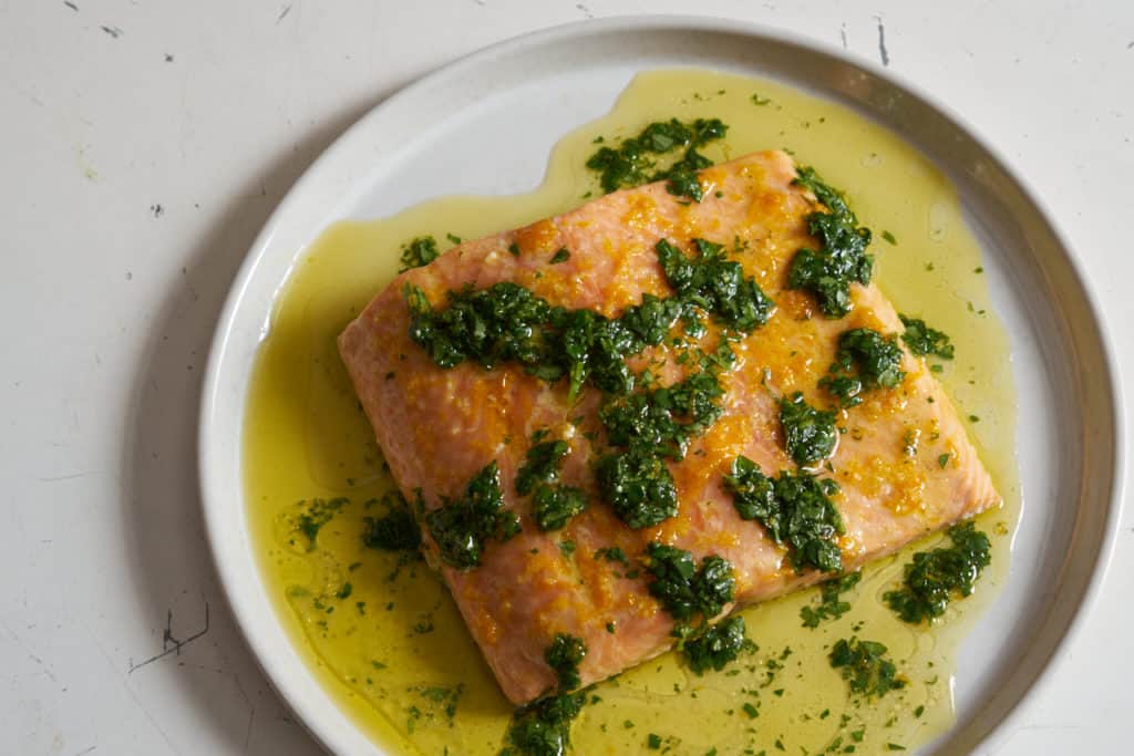 Oven baked salmon with citrus salsa verde on a white plate. Olive oil and herbs surround the salmon filet.