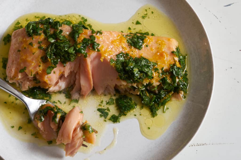 A piece of oven baked salmon with citrus salsa verde on a white plate. A fork is on the left with a bite of salmon on it.