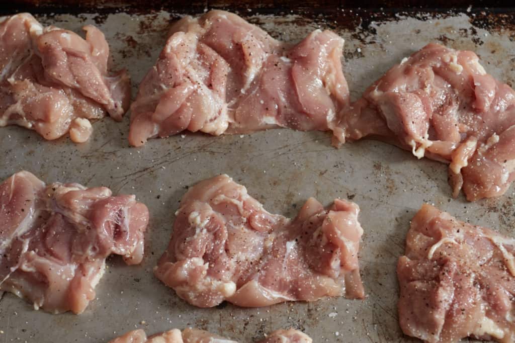 Raw chicken thighs seasoned with salt and pepper on a baking sheet.