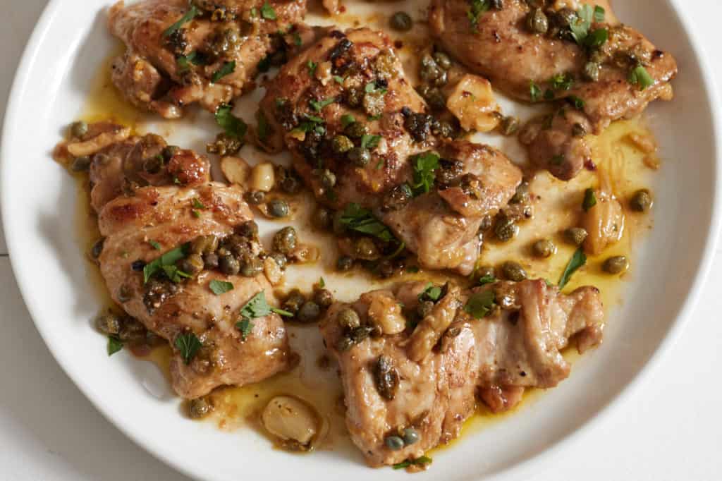 Boneless, skinless chicken thighs with garlic, capers, and lemon topped with fresh parsley on a white plate.