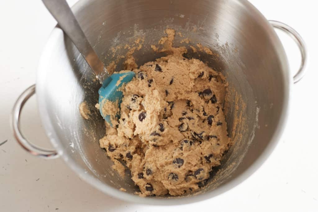 A blue spatula sits in a metal mixing bowl with batter for gluten free chocolate chip cookies.