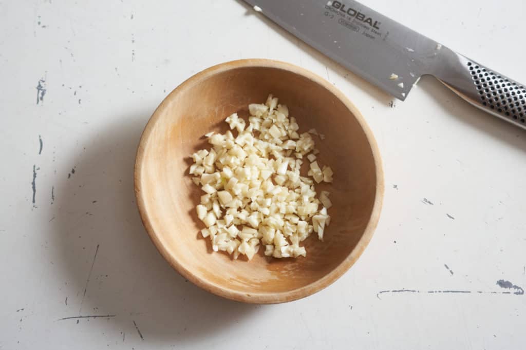 A small wooden bowl with minced garlic next to a stainless steel chef's knife.