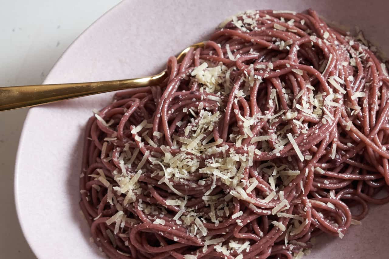 A gold spoon in a pink bowl filled with red wine spaghetti topped with parmigiano cheese.