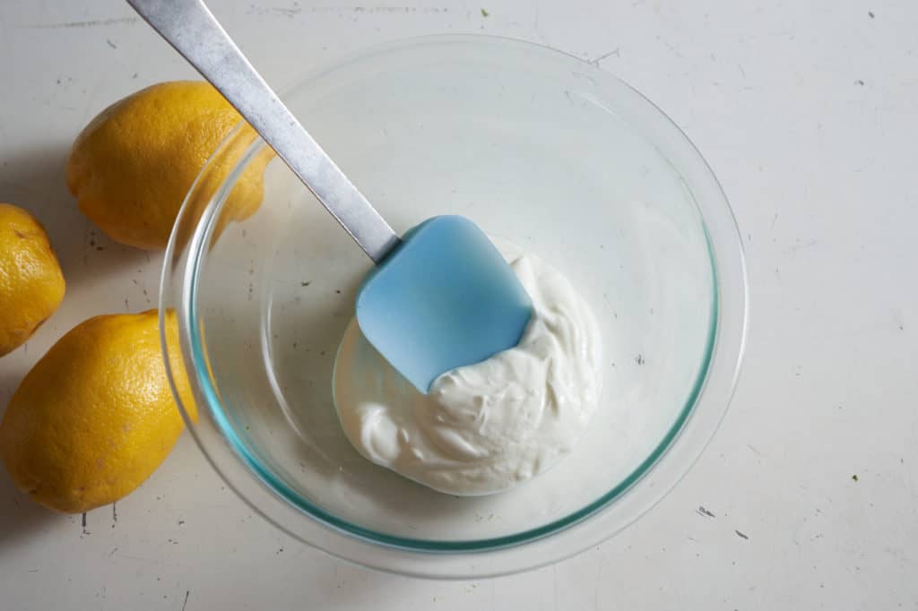 A blue spatula in glass bowl of yogurt sitting next to three lemons.