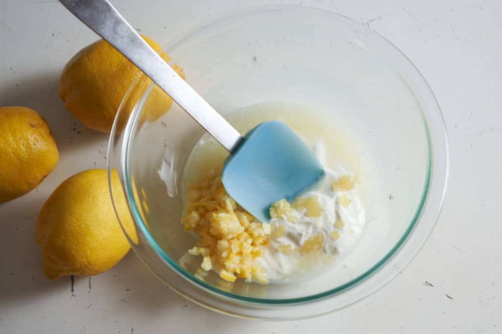 Lemon juice and pieces of lemon peel being stirred into yogurt in a bowl.
