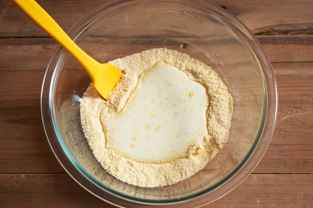 A yellow spatula in a glass bowl with cornmeal and buttermilk.
